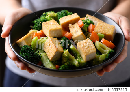 Stir fried tofu, broccoli and carrot in a black bowl holding by woman hand Stir fried tofu, broccoli and carrot in a black bowl holding by woman hand 143557215