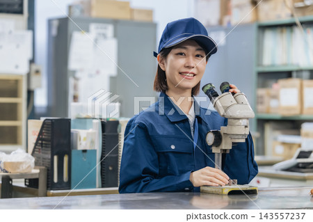 一名工廠工人正在使用顯微鏡。研發、製造、零件、組件、品質控制、檢查、檢驗、驗證、實驗，臉上帶著微笑。 143557237