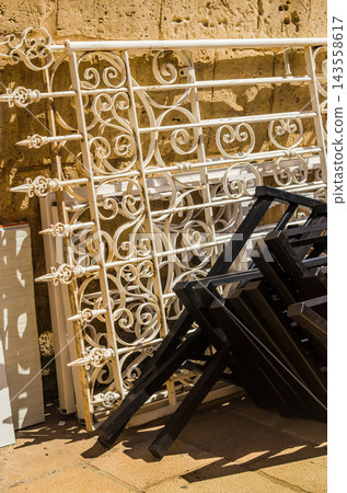A vertical shot showing intricate vintage white metal scrolls and pointed fence spikes stacked alongside folded black furniture frames on stone paving. High quality photo A vertical shot showing intricate vintage white metal scrolls and pointed fence spikes stacked alongside folded black furniture frames on stone paving. High quality photo 143558617