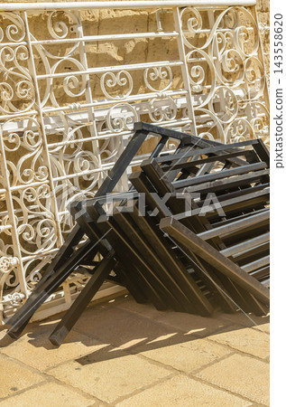A close-up shot of folded black metal furniture frames stacked neatly in front of ornate, vintage white wrought iron balcony railings on a stone pavement.. High quality photo A close-up shot of folded black metal furniture frames stacked neatly in front of ornate, vintage white wrought iron balcony railings on a stone pavement.. High quality photo 143558620