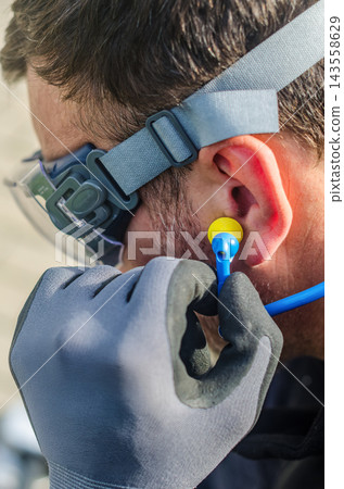 Close-up of man in safety glasses inserting foam earplug for hearing protection, wearing work gloves outdoors. . High quality photo 143558629