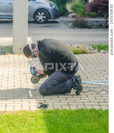 Man wearing safety glasses and gloves kneeling on paved driveway, cutting metal fence rail with angle grinder, sparks flying. High quality photo Man wearing safety glasses and gloves kneeling on paved driveway, cutting metal fence rail with angle grinder, sparks flying. High quality photo 143558630
