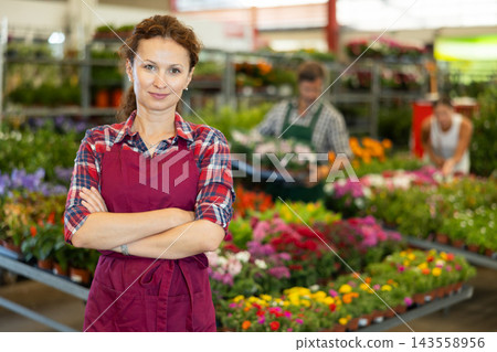 Woman worker stands in aisle between rows with variety of flowering plants in pots Woman worker stands in aisle between rows with variety of flowering plants in pots 143558956