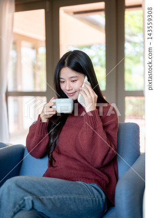 Young Asian woman holding and sipping coffee during smartphone call on sofa in a living room or cafe Young Asian woman holding and sipping coffee during smartphone call on sofa in a living room or cafe 143559308