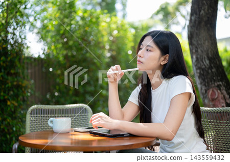 Asian Woman Leaning on Hand Thinking with Stylus at Garden Table Asian Woman Leaning on Hand Thinking with Stylus at Garden Table 143559432