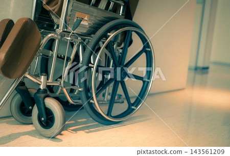 Empty wheelchair near a hospital hallway with black rear wheels, silver hand rims and small front casters, assistive equipment for patient mobility, nursing care and support for older adults. Empty wheelchair near a hospital hallway with black rear wheels, silver hand rims and small front casters, assistive equipment for patient mobility, nursing care and support for older adults. 143561209