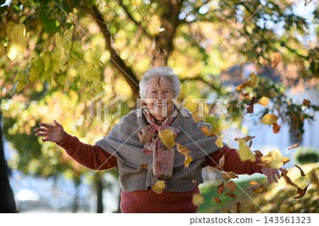 Portrait of elderly woman on walk in the park during warm autumn day, throwing colorful leaves. Portrait of elderly woman on walk in the park during warm autumn day, throwing colorful leaves. 143561323