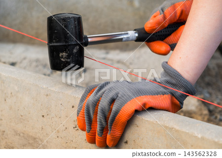 A gloved hand uses a rubber mallet to strike a line on a concrete surface. The precision of construction work, with focus on the tools and materials involved. A gloved hand uses a rubber mallet to strike a line on a concrete surface. The precision of construction work, with focus on the tools and materials involved. 143562328