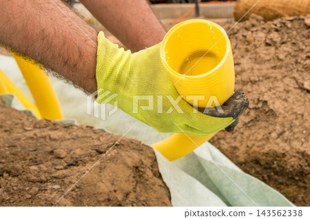 Across a landscaping site, a worker holds a yellow drainage pipe while preparing for installation. The scene includes soil, tools, and equipment. Across a landscaping site, a worker holds a yellow drainage pipe while preparing for installation. The scene includes soil, tools, and equipment. 143562338