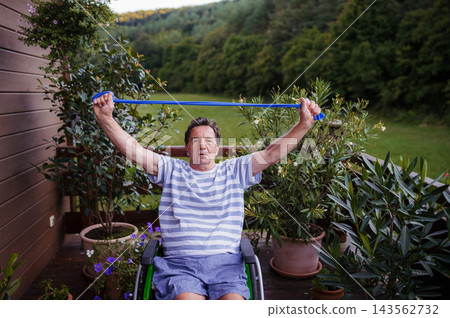 Senior man in wheelchair exercising with resistance band on terrace. 143562732