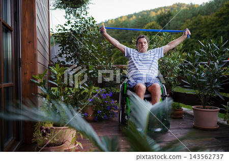 Senior man in wheelchair exercising with resistance band on terrace. Senior man in wheelchair exercising with resistance band on terrace. 143562737