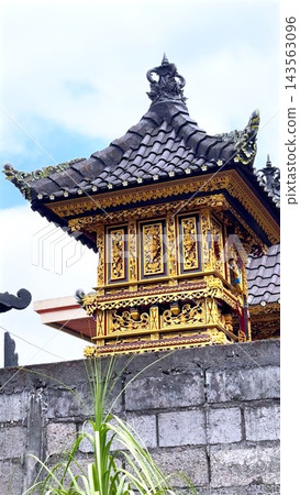 Ornate Balinese temple pavilion stands above stone wall beneath bright sky. Golden floral carvings and dark tiled roof give sacred architecture richly decorative traditional character. Ornate Balinese temple pavilion stands above stone wall beneath bright sky. Golden floral carvings and dark tiled roof give sacred architecture richly decorative traditional character. 143563096