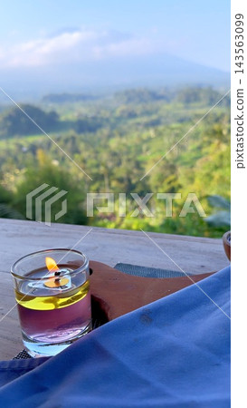 Glass oil lamp burns on wooden terrace overlooking lush tropical hills. Small flame glows beside blue fabric, creating peaceful travel retreat in warm mountain air, while lamp rests on wooden surface. 143563099
