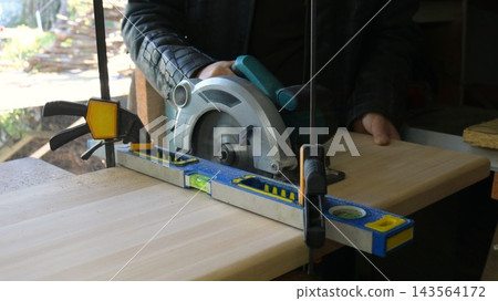 A carpenter in a leather jacket makes a cut with a circular saw, guiding it along a blue-and-yellow level that is rigidly fixed to the wooden tabletop by two quick-release clamps. A carpenter in a leather jacket makes a cut with a circular saw, guiding it along a blue-and-yellow level that is rigidly fixed to the wooden tabletop by two quick-release clamps. 143564172