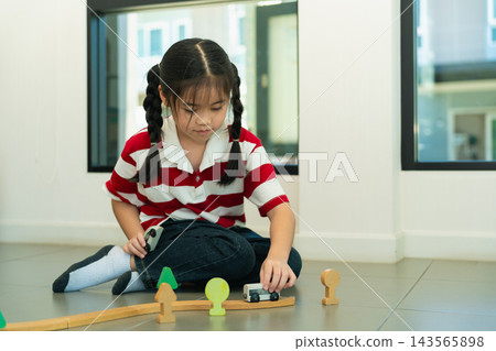 Young Asian girl with braided hair sitting on floor playing with wooden train set and colorful toy trees indoors Young Asian girl with braided hair sitting on floor playing with wooden train set and colorful toy trees indoors 143565898