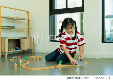 Young girl playing with wooden train track set on tiled floor near window in modern minimalist room interior 143565901