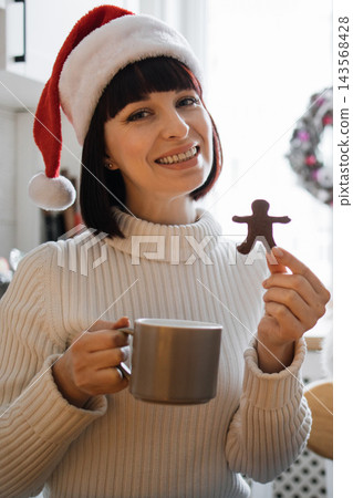 A smiling Caucasian woman wearing a festive Santa hat holds a gingerbread man cookie and a mug of hot beverage A smiling Caucasian woman wearing a festive Santa hat holds a gingerbread man cookie and a mug of hot beverage 143568428
