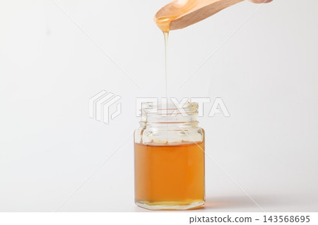 金色的蜂蜜滴入白色背景的玻璃瓶中。 金色的蜂蜜滴入白色背景的玻璃瓶中。 143568695