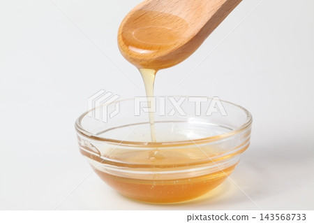 蜂蜜從木勺上滴落到白色背景的玻璃碗中。 蜂蜜從木勺上滴落到白色背景的玻璃碗中。 143568733