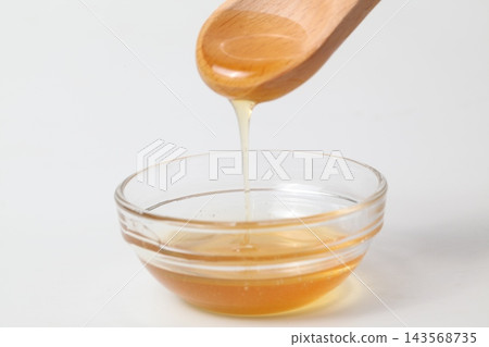 蜂蜜從木勺上滴落到白色背景的玻璃碗中。 蜂蜜從木勺上滴落到白色背景的玻璃碗中。 143568735