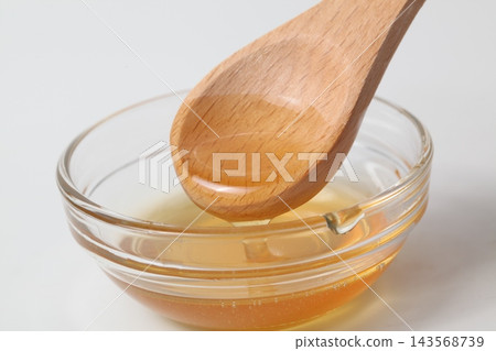 蜂蜜從木勺上滴落到白色背景的玻璃碗中。 蜂蜜從木勺上滴落到白色背景的玻璃碗中。 143568739