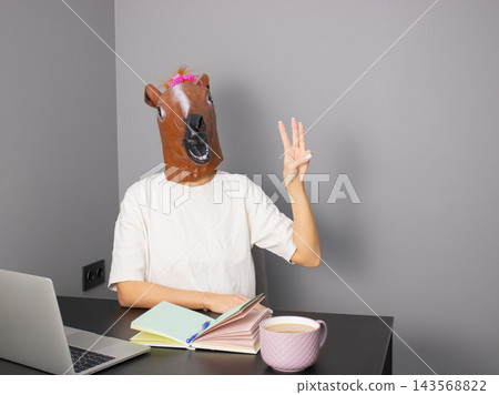 A Humorous Scene Featuring a Person with a Horse Mask Engaging in a Quirky Work Environment with Writing Tools and a Mug A Humorous Scene Featuring a Person with a Horse Mask Engaging in a Quirky Work Environment with Writing Tools and a Mug 143568822