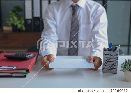 Office worker handing over an envelope, next to a packing box filled with personal belongings. Concept for resignation or leaving a job. Office worker handing over an envelope, next to a packing box filled with personal belongings. Concept for resignation or leaving a job. 143569082