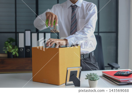 Office worker packing personal belongings into a cardboard box at a desk after job resignation or termination. Office worker packing personal belongings into a cardboard box at a desk after job resignation or termination. 143569088