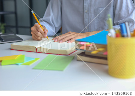University Student Studying and Writhing in Notebook Preparing for Exam with Textbook on the Table at Home. University Student Studying and Writhing in Notebook Preparing for Exam with Textbook on the Table at Home. 143569108