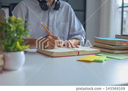 Close-up young man writing in the notebook while studying on the table with laptop and books. 143569109