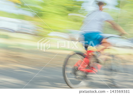 Cyclist Riding Red Bicycle With Motion Blur. Cyclist wearing blue shorts rides red bicycle along sunlit road with strong motion blur. Blurred speed, outdoor activity, dynamic personal transportation 143569381