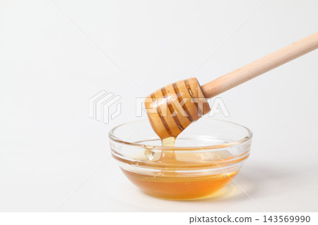 Honey dripping from a honey dipper into a glass dish against a white background. 143569990