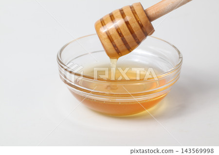 Honey dripping from a honey dipper into a glass dish against a white background. 143569998