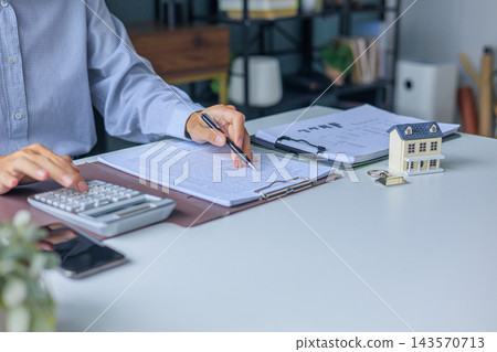 Businessman reading contract document and using calculator next to miniature house on the table in office. Concept for real estate investment, loan, rent, mortgage, insurance Businessman reading contract document and using calculator next to miniature house on the table in office. Concept for real estate investment, loan, rent, mortgage, insurance 143570713