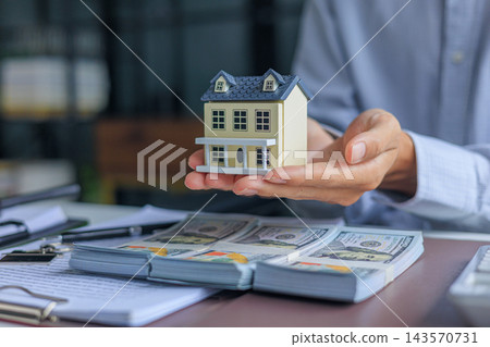 Businessman, Banker, Realtor at office desk holding a house model with money, calculator, and financial documents or contract on the table Businessman, Banker, Realtor at office desk holding a house model with money, calculator, and financial documents or contract on the table 143570731