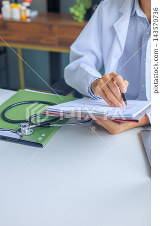 Healthcare professional in a white coat reviewing and filling out patient documentation on a clipboard next to a stethoscope and a laptop Healthcare professional in a white coat reviewing and filling out patient documentation on a clipboard next to a stethoscope and a laptop 143570736