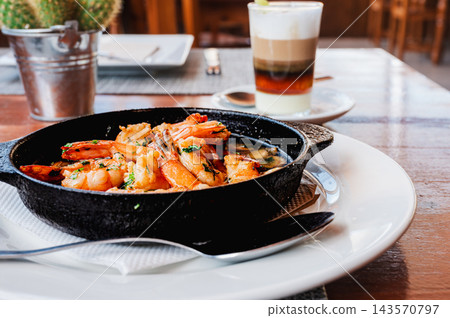 Garlic prawns and coffee on a polished restaurant table. A dish of garlic prawns and a barraquito coffee sit on a polished wooden table in a Canarian restaurant. Relaxed island dining for travel and h Garlic prawns and coffee on a polished restaurant table. A dish of garlic prawns and a barraquito coffee sit on a polished wooden table in a Canarian restaurant. Relaxed island dining for travel and h 143570797