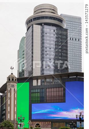 A modern high-rise skyscraper with a round observation deck dominates the skyline above a commercial building featuring large LED screens and a classic clock tower landmark, capturing the dynamic A modern high-rise skyscraper with a round observation deck dominates the skyline above a commercial building featuring large LED screens and a classic clock tower landmark, capturing the dynamic 143571279