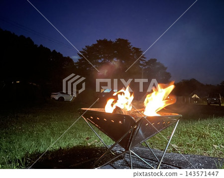 營火在漆黑的夜空下熊熊燃燒,遠處是露營地的景色。 營火在漆黑的夜空下熊熊燃燒,遠處是露營地的景色。 143571447