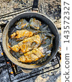 Fried fish is being prepared in a pot over an open fire while camping outdoors. The fish is sizzling in the pan, surrounded by natural elements 143572327
