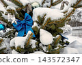A Christmas and New Year tree stands covered in snow, adorned with blue bows and silver ornaments in a winter scene. The tree is outdoors in a snowy 143572347