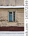 A wall of an old wooden house shows a blue window with cracked glass. Snow covers the ground beneath it. This scene is set in winter 143572349