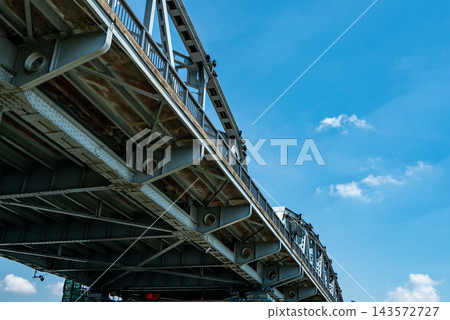 Steel road bridge with riveted girders, diagonal bracing and metal railings beneath blue sky and white clouds, weathered concrete decking supported by a truss structure for transport infrastructure. 143572727