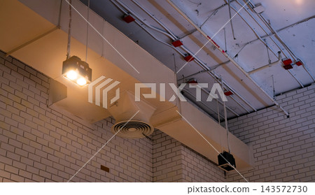 Ceiling lamps suspended beside exposed air conditioning ducts and a circular diffuser, fire sprinkler pipes and electrical conduits beneath concrete ceiling above white brick walls inside a building. Ceiling lamps suspended beside exposed air conditioning ducts and a circular diffuser, fire sprinkler pipes and electrical conduits beneath concrete ceiling above white brick walls inside a building. 143572730