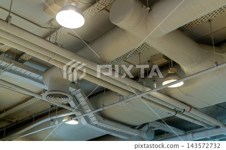 Air ducts, wiring and plumbing exposed on a shopping mall ceiling, circular ventilation diffusers and hanging lamps among white pipes and cable trays, mechanical and electrical building services. 143572732
