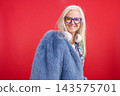 Senior woman smiling, wearing a blue fur coat, headphones, and glasses 143575701