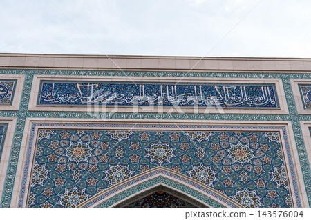 Ornate turquoise and blue mosaic tiles surround Arabic calligraphy and a pointed arch on a richly patterned Islamic architectural facade beneath a pale sky Ornate turquoise and blue mosaic tiles surround Arabic calligraphy and a pointed arch on a richly patterned Islamic architectural facade beneath a pale sky 143576004