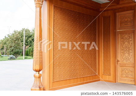 Detailed carved wood entrance features geometric panels, a decorative door, and column beside a paved outdoor area with trees and greenery Detailed carved wood entrance features geometric panels, a decorative door, and column beside a paved outdoor area with trees and greenery 143576005