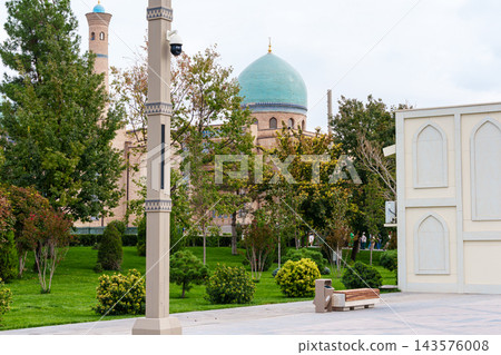 Turquoise dome and slender minarets rise behind leafy trees, manicured shrubs, benches, and a decorative facade along a paved courtyard Turquoise dome and slender minarets rise behind leafy trees, manicured shrubs, benches, and a decorative facade along a paved courtyard 143576008