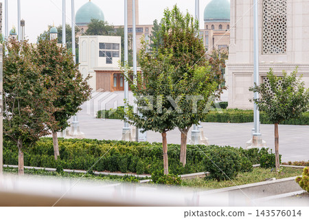 Manicured courtyard with small trees, hedges, white columns, ornate latticework, and turquoise domes seen across a paved architectural complex Manicured courtyard with small trees, hedges, white columns, ornate latticework, and turquoise domes seen across a paved architectural complex 143576014